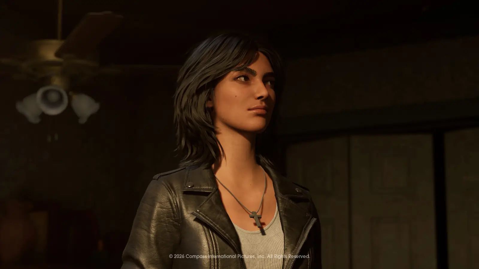 A dark-haired woman in a black leather jacket over a pale top, wearing a cross pendant, stands indoors under a ceiling fan and looks off to her left.