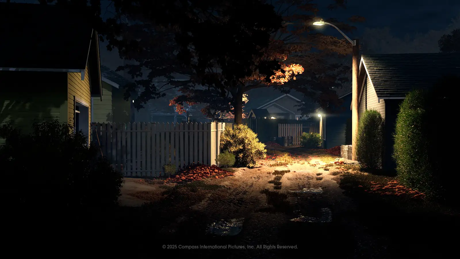 A lamplit path between houses and white picket fences at night, with autumn leaves and a large tree in the center.